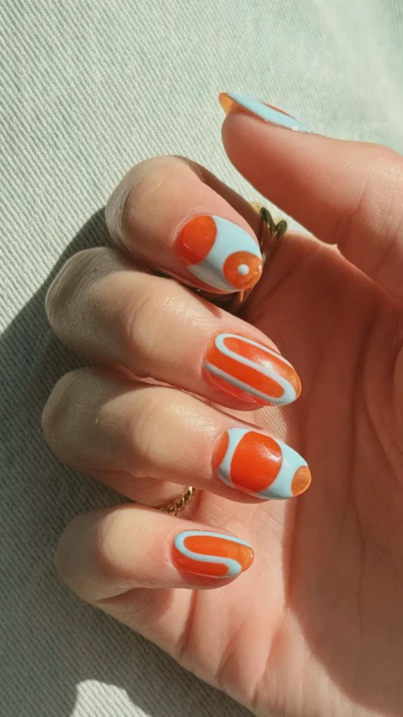 A close-up of a hand showcasing a modern, short almond nail art design. The manicure features a chic, retro-inspired color palette of vibrant burnt orange and pale pastel blue. Each nail has a unique abstract geometric pattern: some have smooth, flowing swirls, while others feature blocky negative space or dot accents. The look combines a glossy finish with clean lines, styled with gold rings against a softly lit, textured fabric background