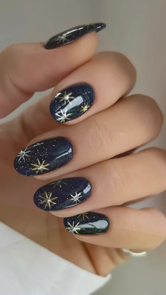 A close-up of a hand showcasing a winter-themed manicure. The nails are shaped in a smooth, glossy oval and feature a deep, midnight blue base coat with a fine silver glitter overlay, resembling a starry night sky. Hand-painted, multi-pointed starbursts and celestial snowflakes in metallic silver and gold are delicately detailed across each nail, creating a cozy and magical winter aesthetic.