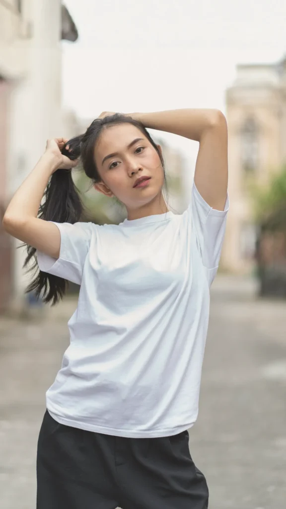 A young woman posing outdoors in a clean, minimalist outfit consisting of a crisp white t-shirt and black tailored trousers. She is adjusting her long dark hair into a ponytail against a blurred urban background.