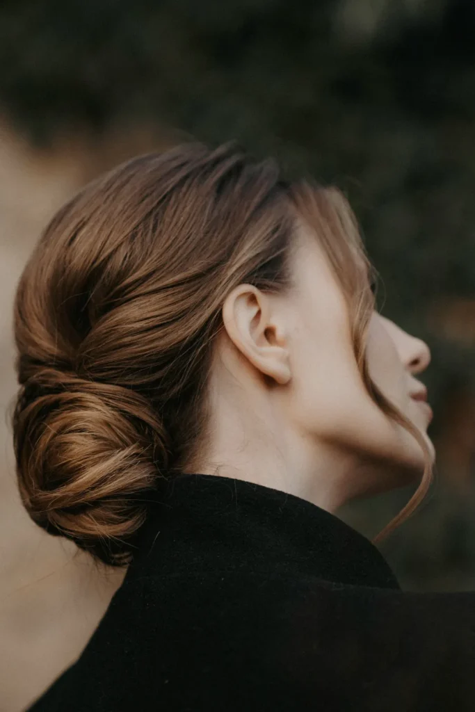 A close-up side view of a polished low bun hairstyle on a woman with warm brown hair. The hair is smoothly swept back into a neat twist at the base of the head, with a soft tendril framing her face. She is wearing a black blazer or coat.