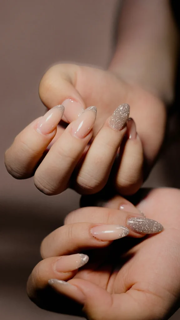 A close-up of a person's hands showcasing a short almond-shaped manicure. The nails feature a mix of neutral designs, including classic French tips with a shimmering silver glitter finish and full-coverage champagne glitter accent nails, set against a soft, blurred background.