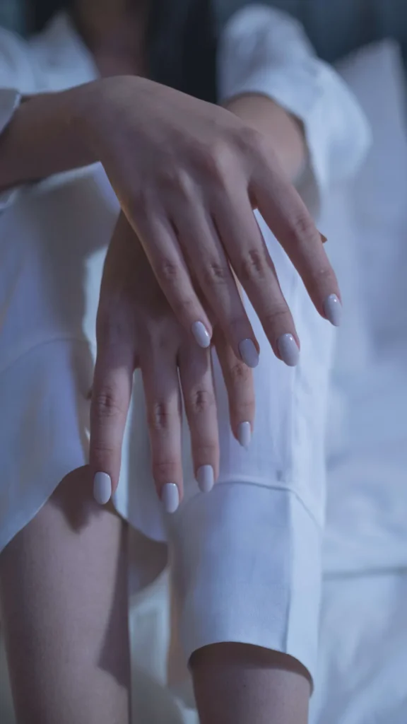 Close-up of a person's hands resting elegantly on their lap, showcasing a clean, minimalist manicure in a milky white or soft neutral shade. The nails are shaped in a medium-length almond style with a glossy finish, epitomizing the "quiet luxury" aesthetic. The person is wearing a crisp white outfit, and the soft, natural lighting emphasizes a polished, sophisticated, and timeless look.