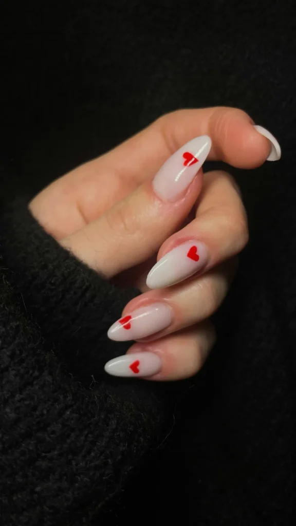 A close-up of a hand featuring an elegant manicure on long, slender almond-shaped nails. The base color is a sheer, high-gloss milky white polish that looks clean and translucent. Each nail is accented with a tiny, vibrant red heart painted near the tip or center, adding a minimalist and playful touch. The hand is posed gently against a cozy, dark black knit sweater background.