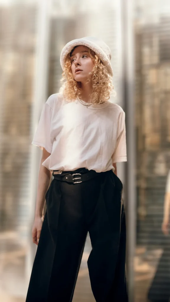 A woman with blonde curly hair wearing a minimalist, expensive-looking outfit featuring a thick cream t-shirt tucked into navy blue tailored trousers. She is accessorized with a black leather belt, a pearl necklace, and a white fuzzy bucket hat, standing against a blurred architectural background.