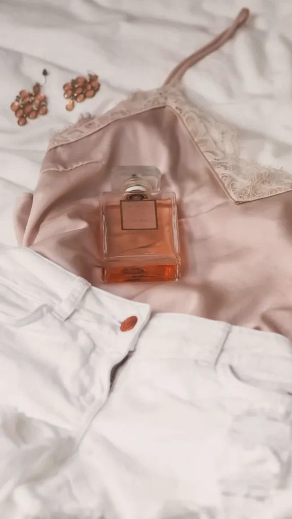 A flat lay of luxury fashion essentials featuring a champagne-colored silk camisole with lace trim, white denim shorts, a bottle of designer perfume, and elegant rose-gold statement earrings resting on a white textured surface.