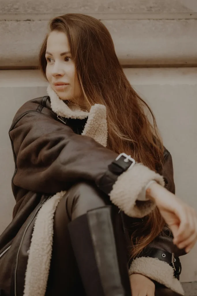 A young woman with long, voluminous brunette hair styled in a '90s-inspired blowout, sitting outdoors in a dark brown shearling-lined aviator jacket and leather trousers. Her hair is full of body and layered, cascading over her shoulder.