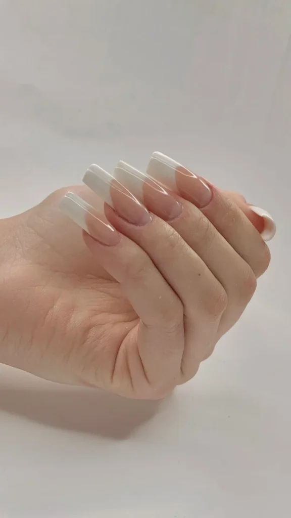 A close-up of a hand featuring a classic French manicure on long, square-shaped nails. The design includes a sheer nude base with crisp, thick white tips, set against a plain, neutral background.