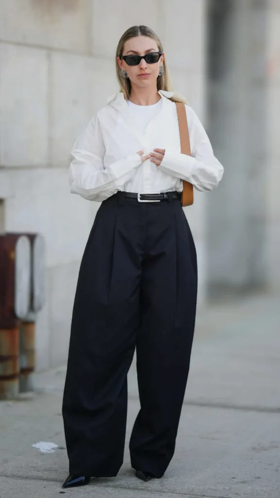 A full-length shot of a woman showcasing a classic, elevated outfit on a city sidewalk. She is wearing high-waisted, wide-leg pleated black trousers secured with a thin black belt featuring a silver buckle. Tucked into the trousers is a crisp, oversized white button-down shirt layered over a simple white crewneck undershirt. She accessorizes the timeless look with black pointed-toe shoes, sleek black sunglasses, chunky silver earrings, and a tan leather shoulder bag, perfectly illustrating the "quiet luxury" aesthetic.