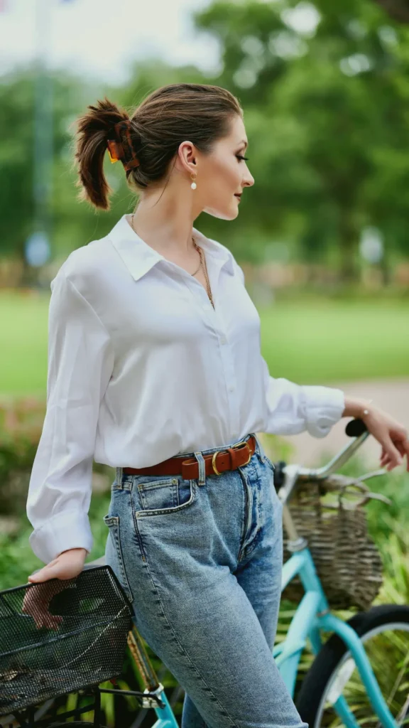 A woman standing in a lush green park leaning against a light blue bicycle. She is wearing a crisp, white button-up shirt tucked into high-waisted denim jeans with a brown leather belt. Her hair is pulled back in a tortoiseshell claw clip, and she is wearing elegant gold pearl earrings, embodying a "quiet luxury" weekend aesthetic.
