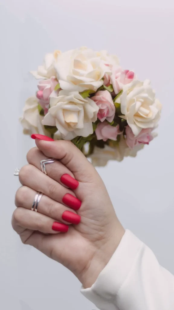 A hand holding a small bouquet of white and pink roses, featuring a vibrant red matte manicure. The nails are shaped in a medium-length square style with a velvety, non-glossy finish. The hand is adorned with simple silver rings and is wearing a white long-sleeved garment against a neutral background.