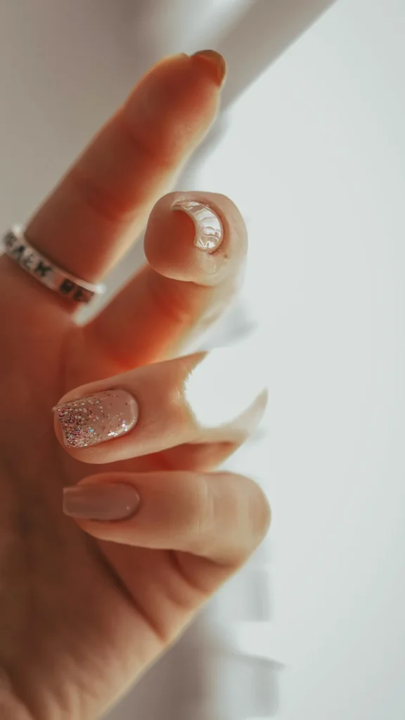 A close-up of a hand showcasing a glossy neutral nude manicure on short, squoval-shaped nails. The ring finger features a subtle accent of fine pink and silver glitter concentrated at the tip. The thumb displays a shiny, metallic silver finish. The hand is softly lit, wearing a silver band ring, set against a bright, minimalist white background.