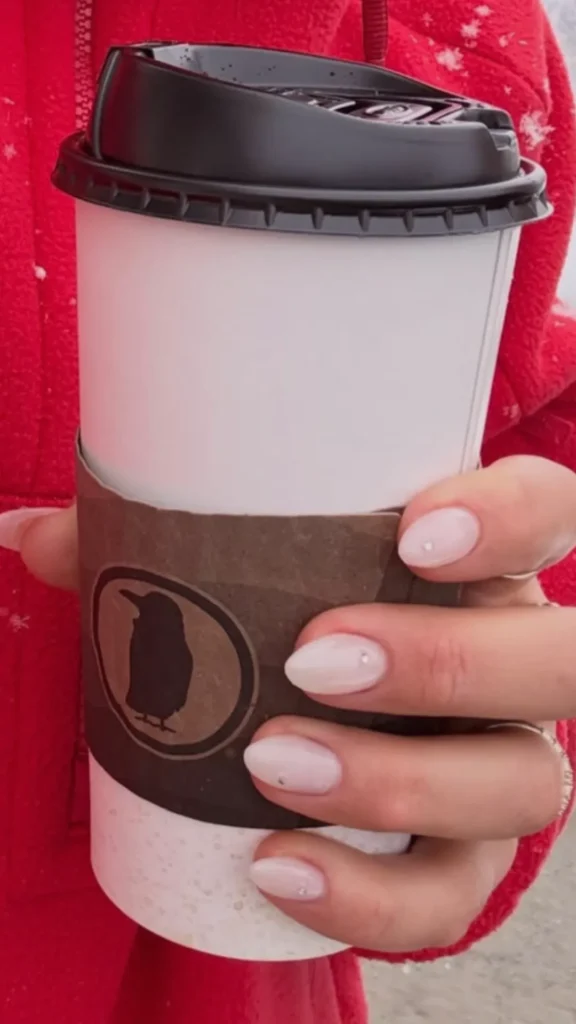A close-up shot focusing on a woman's manicured hand holding a white takeaway coffee cup with a brown cardboard sleeve featuring a penguin logo. She is wearing a bright red fleece jacket. Her nails are shaped into a sophisticated, soft almond style and painted with a clean, milky-white neutral polish. Each nail is subtly accented with a tiny, single silver rhinestone or dot detail near the base, exemplifying a neat, intentional, and polished grooming routine.