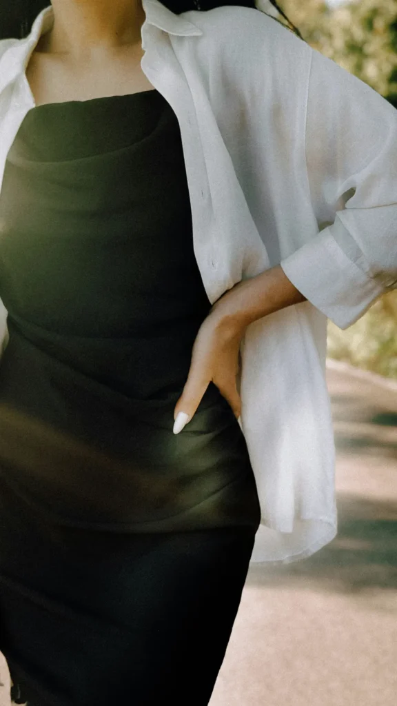 A close-up of a woman wearing a charcoal gray slip dress made of smooth, slightly shimmering fabric. She has layered a crisp white button-up shirt over the dress for a relaxed, expensive-looking minimalist aesthetic. One hand is resting on her hip, showcasing the drape of the fabric and her white manicured nails.