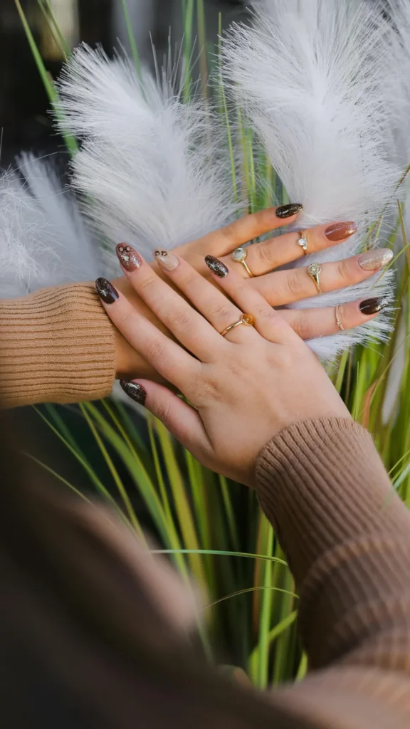 Two hands are crossed gently over a backdrop of fluffy white pampas grass and green stems, showcasing a multi-colored autumn-inspired manicure on almond-shaped nails. The nail design features a mix of dark chocolate brown with fine glitter, solid warm tan, a light beige nail with subtle animal print accents, and champagne shimmer. The person is wearing a cozy, ribbed taupe sweater and several delicate gold rings.
