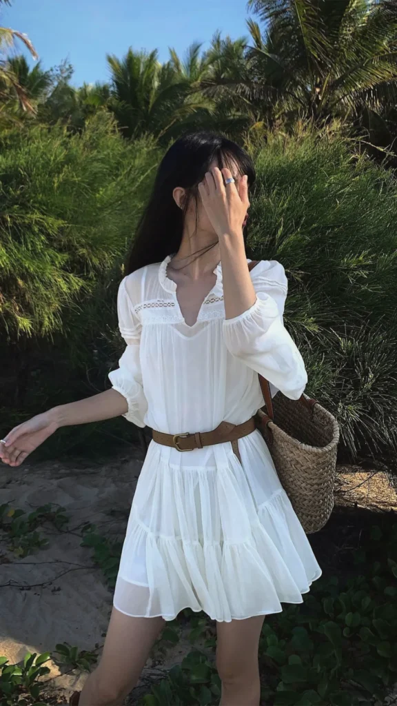 A woman stands outdoors in front of lush tropical greenery, showcasing an effortless Parisian-inspired outfit. She is wearing a breezy white bohemian-style mini dress with sheer half-sleeves and delicate lace detailing at the neckline, accessorized with a rugged brown leather belt cinched around her waist. She carries a classic woven straw tote bag over her shoulder, capturing the perfect blend of natural comfort and quiet luxury style.
