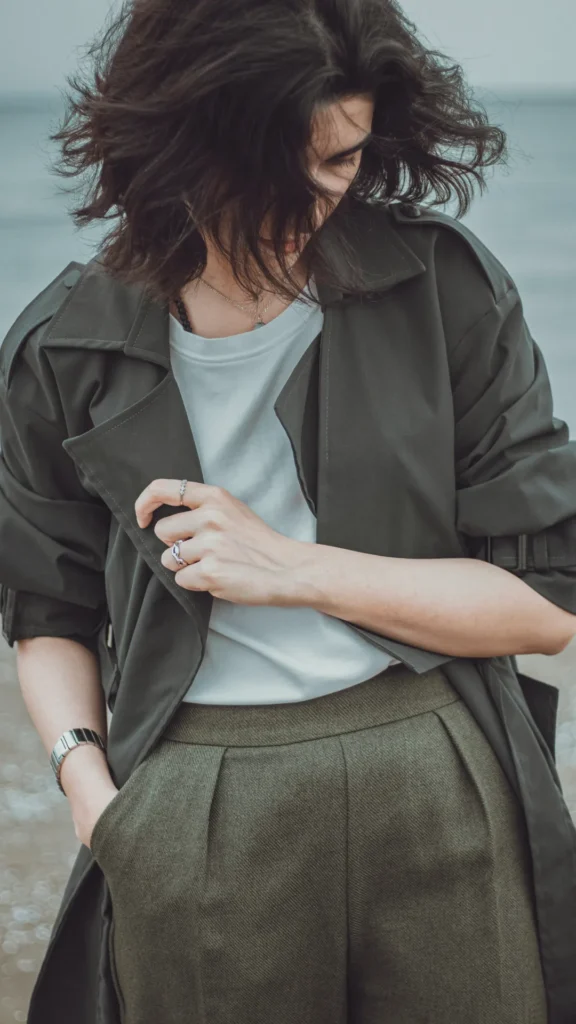 A close-up of a woman wearing an effortlessly chic summer outfit. She is wearing high-waisted, olive-green textured trousers and a light grey crew-neck t-shirt, layered under a matching olive-drab lightweight trench coat or overshirt with rolled sleeves. The look is accessorized with silver rings, a delicate necklace, and a metal watch.