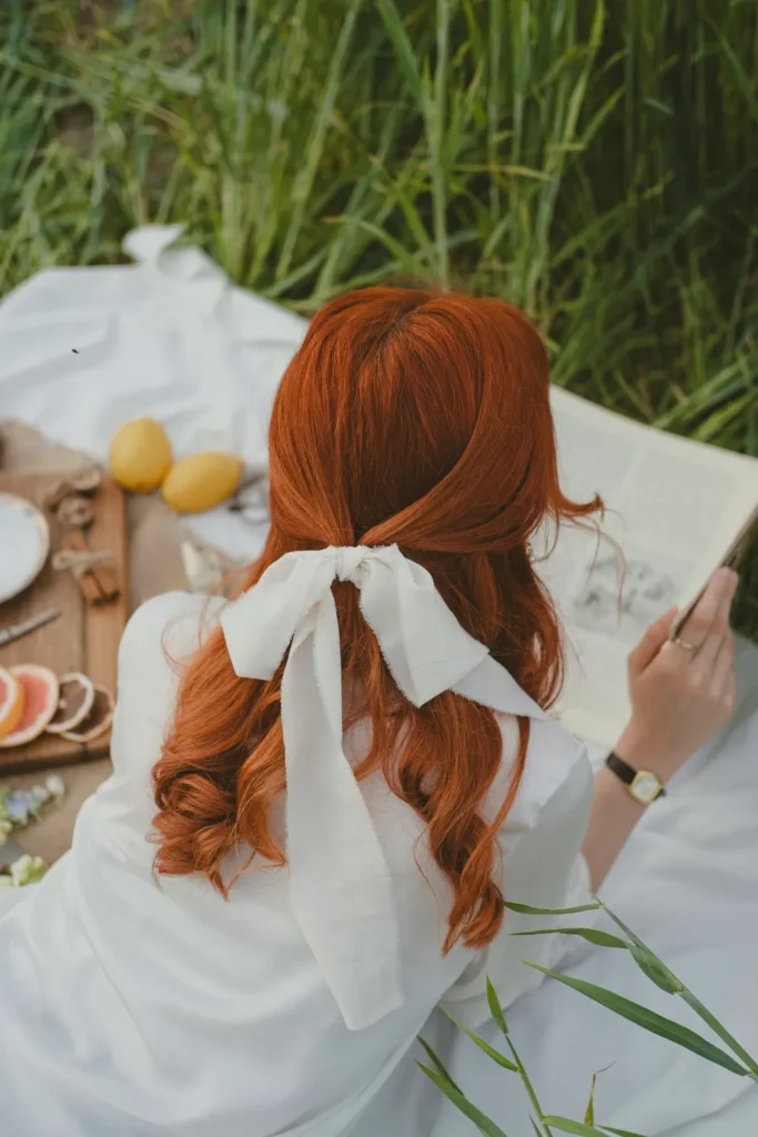 Rear view of a woman with vibrant red hair styled in half-up long hairstyles for the quiet luxury aesthetic, secured with a delicate white silk ribbon during a minimalist outdoor picnic.