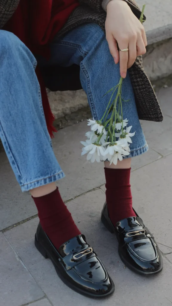 Close-up of a person sitting outdoors wearing polished black patent leather loafers with gold hardware, paired with deep burgundy socks and straight-leg blue jeans. They are holding a small bouquet of white daisies against their leg, capturing a classic "Old Money" French aesthetic.