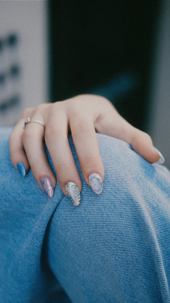 A hand with medium-length almond nails resting on blue denim jeans. Each nail features a unique, 3D textured design with a mix of matte and metallic finishes in shades of blue, silver, and iridescent lavender. The intricate art includes raised swirl patterns and abstract shapes, giving the manicure a modern, edgy, and high-fashion aesthetic.