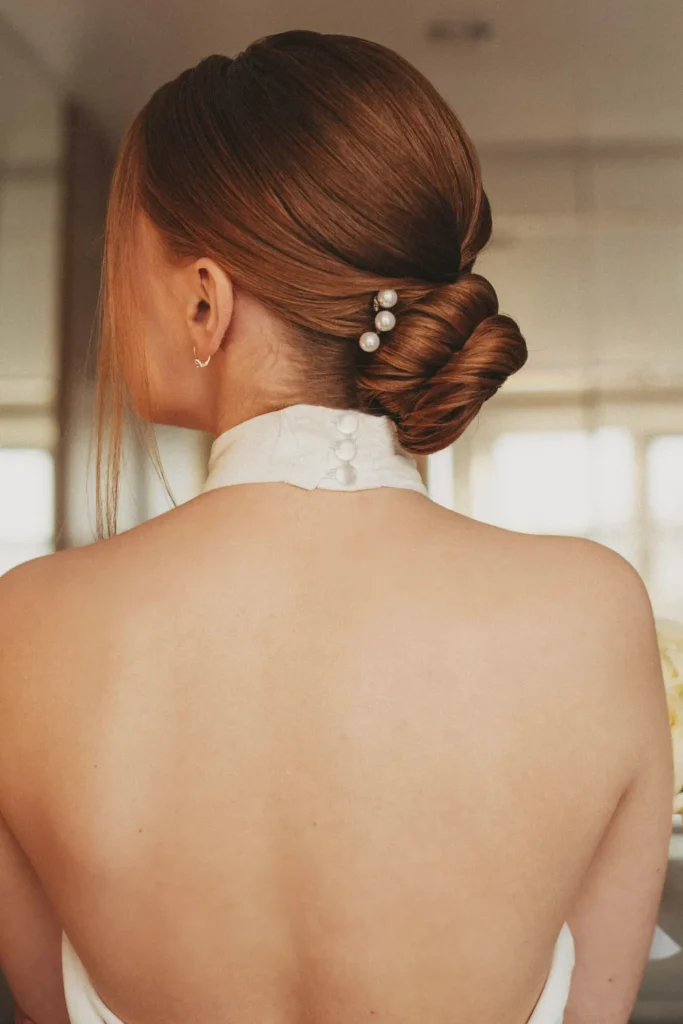 A rear view of a woman with sleek, reddish-brown hair styled in a polished low bun at the nape of the neck. Three decorative pearl pins are tucked into the side of the twist, and she is wearing a high-collared white dress with button details, creating an elegant and sophisticated aesthetic.