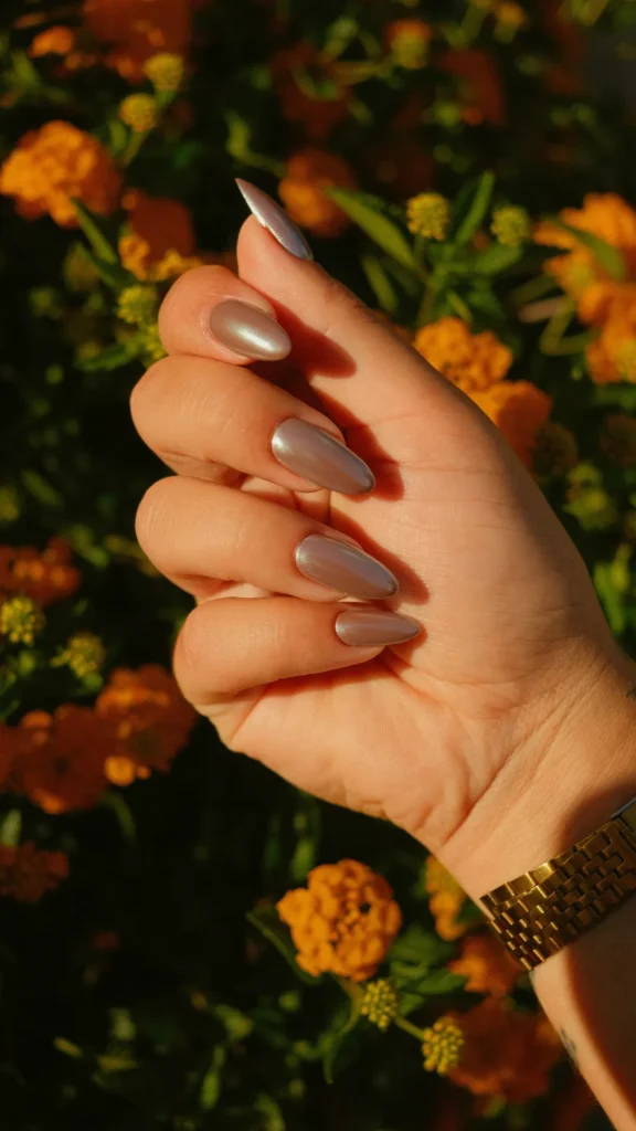 A hand displays a glossy, metallic chrome manicure on long, slender almond-shaped nails. The polish is a shimmering, reflective taupe shade with a silvery undertone. The hand is bathed in warm golden hour sunlight, wearing a gold link watch band, with a vibrant background of blooming orange and yellow lantana flowers.
