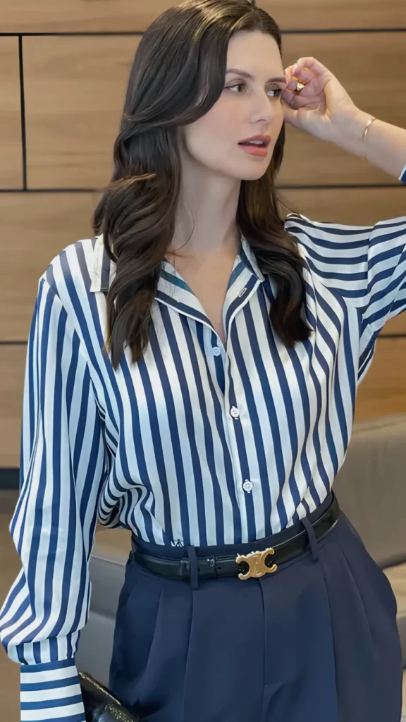 A close-up of a woman modeling a sophisticated, polished outfit that embodies quiet luxury. She is wearing a silk, long-sleeved button-down shirt with vertical navy blue and white stripes, neatly tucked into tailored high-waisted navy trousers. The look is elevated with a black leather belt featuring a prominent gold Celine logo buckle, showcasing how natural fabrics and precise tailoring create effortless comfort and confidence.