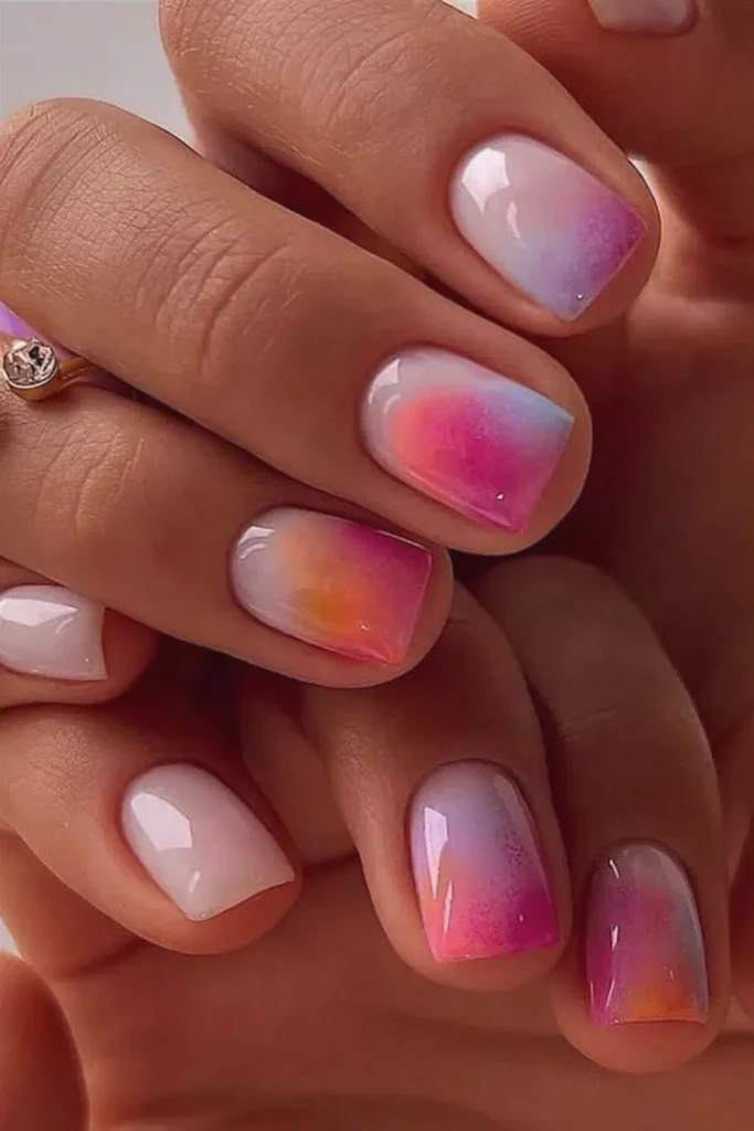 Close-up of hands showing a short, square-shaped manicure with a glossy finish. The nails feature a soft, milky-white base that beautifully blends into a vibrant, airbrushed aura ombré effect at the tips. The colorful gradient mimics a twilight sunset, mixing shades of magenta pink, warm orange, soft purple, and a hint of pastel blue for a dreamy, sunset-sky appearance.