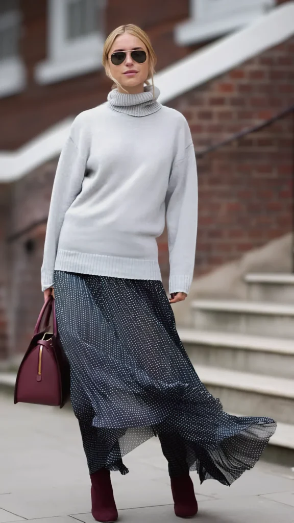 A full-length street style shot of a woman walking near outdoor stone steps, showcasing a masterclass in mixing textures. She is wearing an oversized, cozy light-gray knit turtleneck sweater layered over a flowing, lightweight navy blue pleated maxi skirt adorned with small white polka dots. Her accessories feature rich, matte finishes, including a structured deep burgundy leather handbag and matching burgundy suede ankle boots. She completes the effortless, upscale look with classic aviator sunglasses and her blonde hair tied back, leaving a few loose strands to frame her face