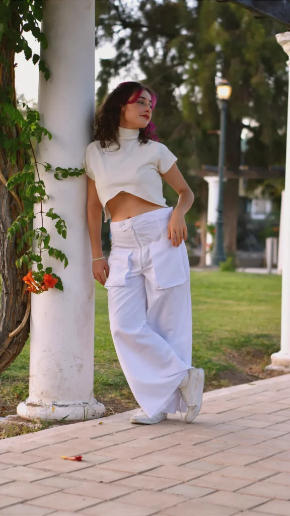 A young woman leaning against a white architectural pillar in a park, wearing an all-white monochromatic outfit. The look features a high-neck cropped cream t-shirt and oversized, wide-legged white cargo trousers with large pockets, paired with white sneakers. Her hair has vibrant pink highlights, adding a pop of color to the clean, minimalist aesthetic.