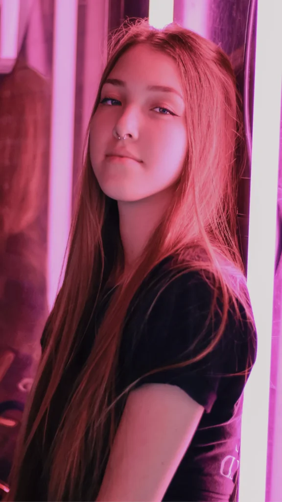 A young woman with long, straight hair and a natural, glowing complexion leans against a wall illuminated by pink neon light in 6.webp. She wears minimal makeup with a subtle winged eyeliner and a septum piercing, embodying an effortless and "undone" beauty aesthetic.