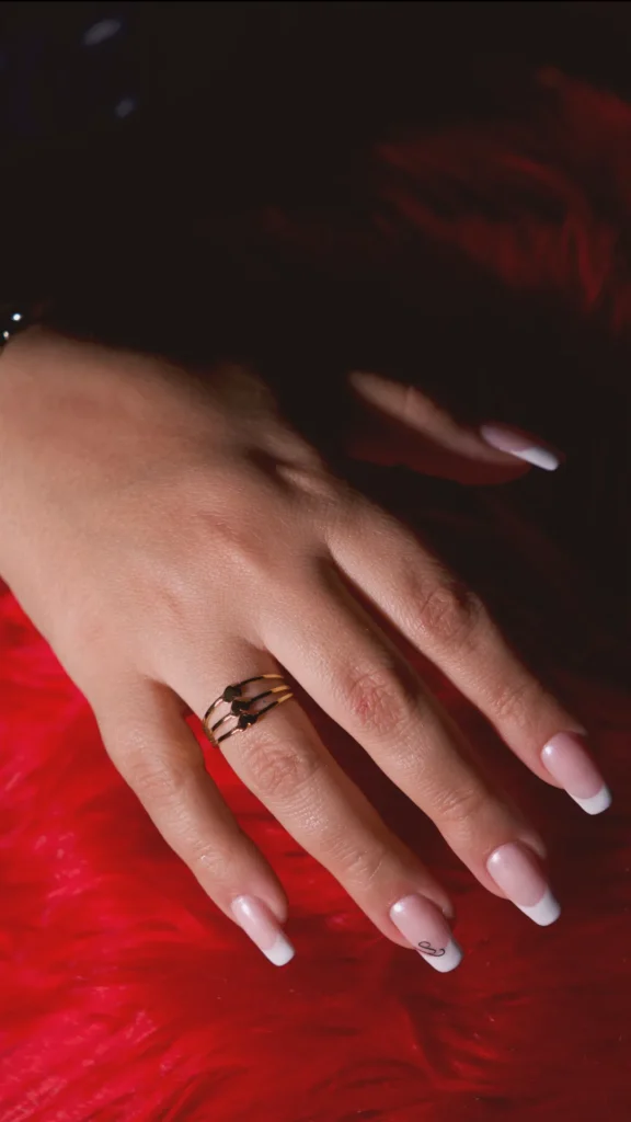 A hand with a classic French manicure featuring crisp white tips on a natural pink base. The ring finger is styled with a delicate triple-band gold ring, and the pinky nail includes a tiny black scrollwork detail. The hand is set against a vibrant, textured red faux-fur background, emphasizing a polished and accessorized look.