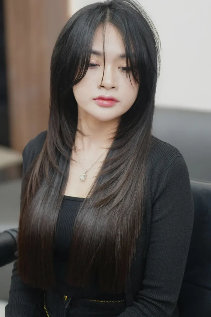 A young woman with long, straight black hair styled in a sleek "glass hair" finish with subtle layers and face-framing bangs. She is wearing a monochromatic black outfit consisting of a ribbed cardigan and top, paired with a simple silver pendant necklace.