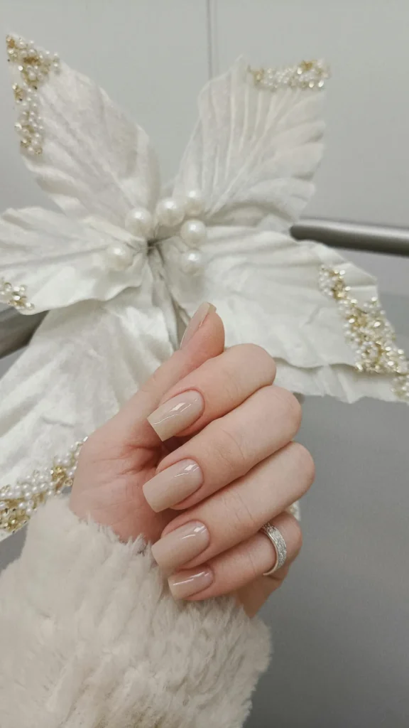 A close-up of a hand showcasing a classic, high-gloss nude manicure on medium-length, square-shaped nails. The polish is a clean, semi-sheer beige that perfectly matches the skin's natural undertones. The hand is wearing a textured, fluffy white winter sweater and a sparkling silver pavé band ring, holding a white decorative holiday flower with faux pearls and gold beads.