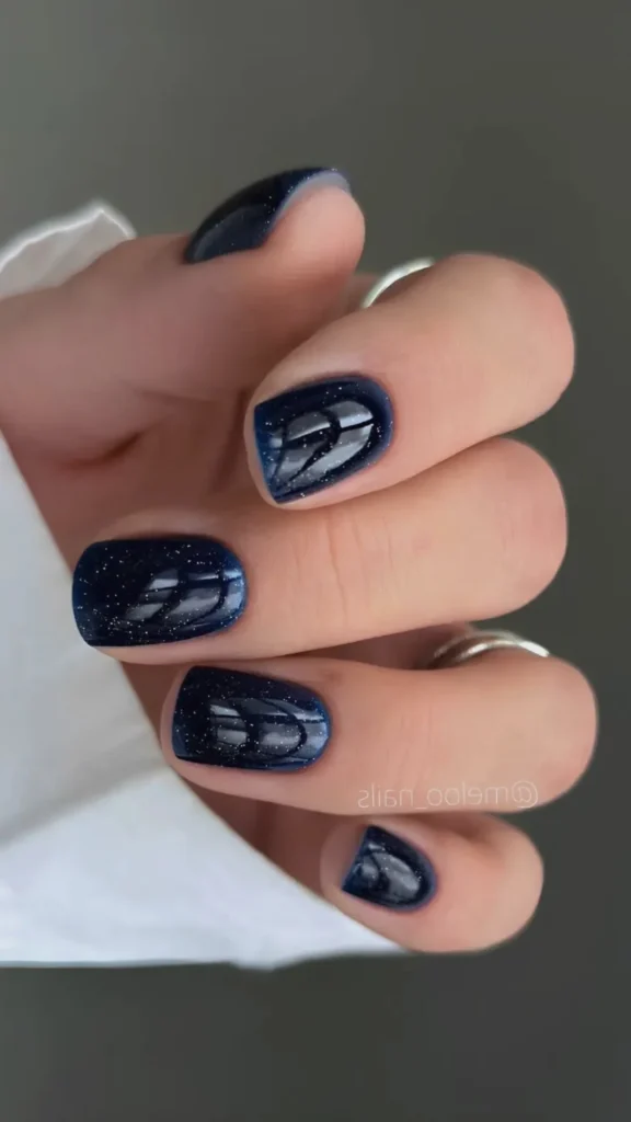 A close-up of a hand showcasing a glossy winter manicure on short, squoval-shaped nails. The nails feature a deep, high-shine midnight blue base coat dusted with an ultrafine silver glitter that creates a starry, celestial effect. The individual is wearing a minimalist silver ring on their index finger and a stacked set of thin silver bands on their ring finger, beautifully complementing the cool tones of the nail polish.