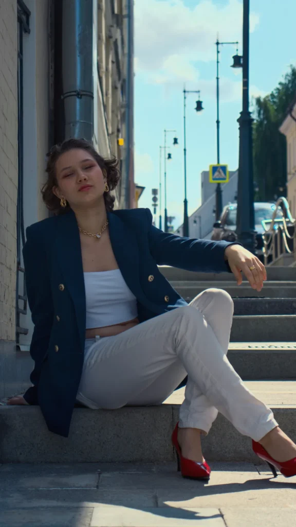 A young woman sitting on stone city steps, wearing a structured navy blue double-breasted blazer with gold buttons over a white cropped top and white slim-fit trousers. She is wearing bold red high heels and chunky gold jewelry, including a chain necklace and hoop earrings, embodying a polished "quiet luxury" aesthetic.