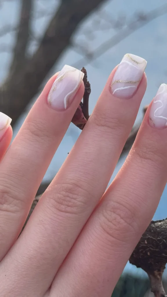 A close-up view of fingers showcasing a manicure on long, square-shaped nails. The base is a milky, semi-translucent pinkish-white polish. The nail art features delicate, organic abstract swirls painted in white and a textured, glittery gold across the surface of each nail. The hand is positioned outdoors near tree branches, with a soft-focus background of blue sky.