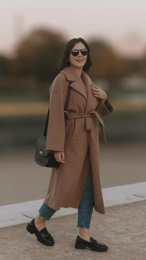 A woman smiling while walking outdoors along a waterfront path, modeling a universally flattering quiet luxury look. She is wearing a classic, long camel-colored wool wrap coat cinched at the waist with a matching belt, layered over straight-leg blue jeans. Her look is accessorized with black chunky leather loafers, a structured black leather shoulder bag, and dark sunglasses, perfectly demonstrating a timeless and elegant outfit style for any age.