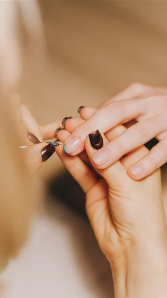 A close-up, top-down view of a professional manicure in progress. A nail technician uses a fine-detail brush to apply a thin turquoise line to the tip of a client's natural-looking almond nail, creating a delicate French tip effect. The technician’s own nails are painted a glossy dark espresso brown, providing a professional contrast as they carefully steady the client's hand.