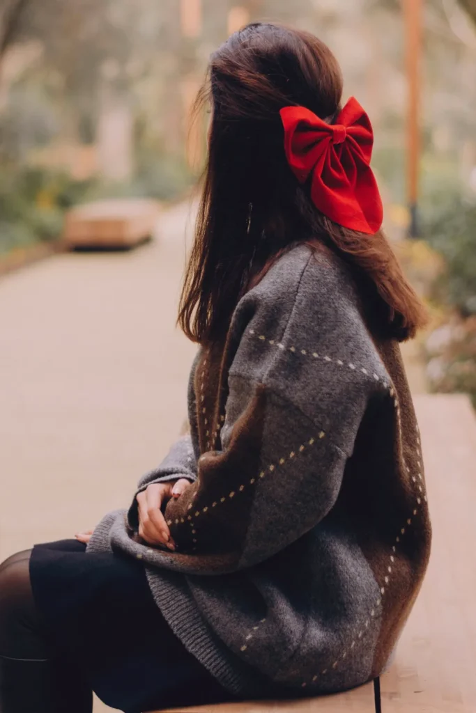 A side-rear view of a woman sitting on a bench in an outdoor park setting during autumn. She has long brunette hair styled in a half-up ponytail, adorned with a large, vibrant red velvet bow. She is wearing an oversized grey and brown patterned wool sweater and a dark skirt, epitomizing a cozy, "preppy" aesthetic.