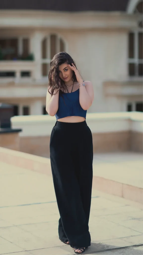 A woman posing outdoors wearing high-waisted, black wide-leg trousers paired with a cropped, ribbed navy blue top. She has long, dark wavy hair and is touching her head with both hands while looking toward the camera. The background shows a blurred, light-colored building terrace.