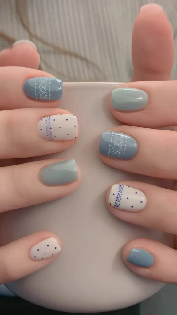 A close-up view of two hands cupping a smooth, matte light grey mug. The short, square-shaped nails feature a glossy, cozy winter sweater nail art design. The color palette alternates between a dusty denim blue, a muted sage green, and a milky off-white base coat. The accent nails are decorated with delicate, hand-painted sweater-knit patterns—including zig-zag lines, cross-hatches, and tiny blue polka dots—evoking a warm, hygge aesthetic.