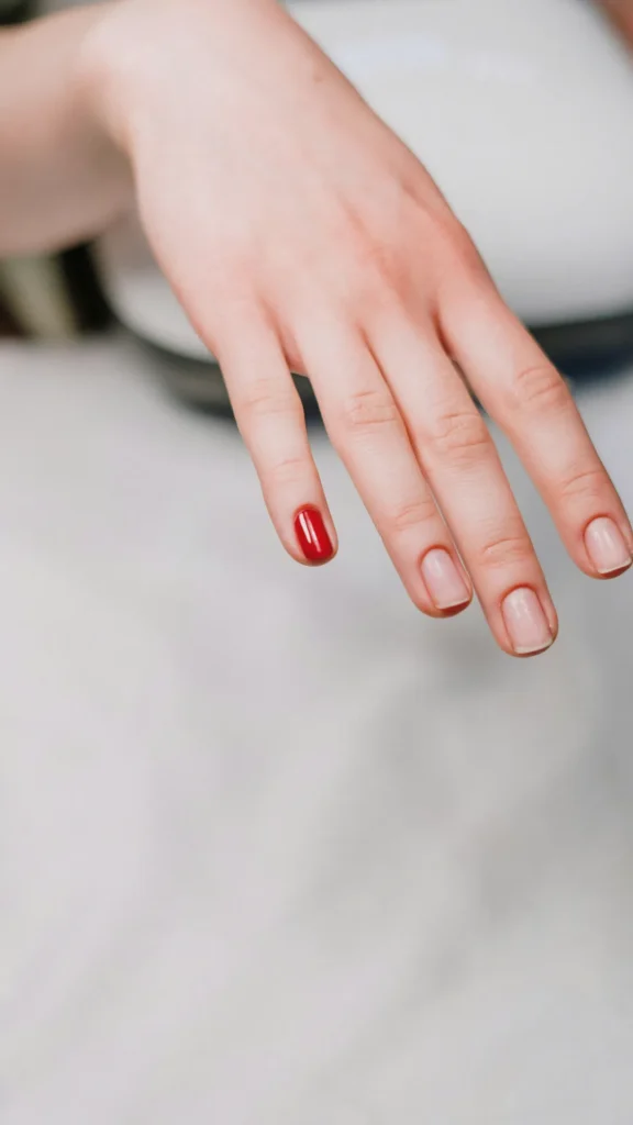 A high-angle view of a person's hand resting over a white surface during a manicure session. The short, naturally rounded pinky nail is painted with a high-gloss, classic bright red nail polish. The remaining three fingers feature clean, unpolished natural nails, capturing a work-in-progress look.