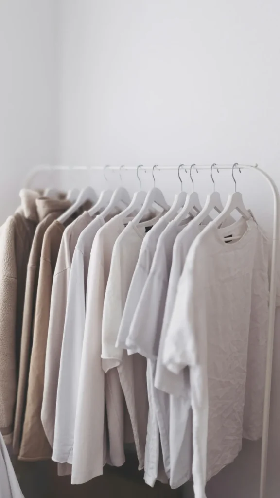 A minimalist 10-piece capsule wardrobe displayed on a white clothing rack against a clean white wall. The collection features a curated selection of sweaters, t-shirts, and light jackets in a cohesive neutral palette of cream, taupe, and soft grey, hanging on uniform white wooden hangers to demonstrate an organized and "expensive-looking" closet system.
