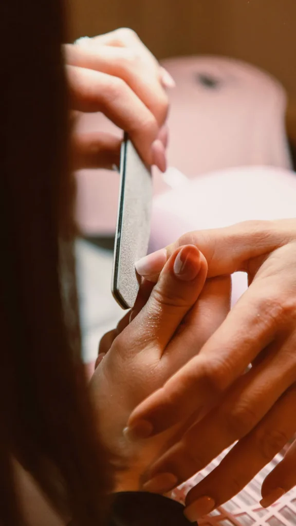 An over-the-shoulder, close-up shot of a professional manicure session in progress. A nail technician is carefully using a grey emery board nail file to shape the edge of a client's fingernail. The client’s nails are painted in a soft, glossy sheer pink shade. The scene is warmly lit with a soft-focus background, highlighting the careful, intentional process of nail care and maintenance.