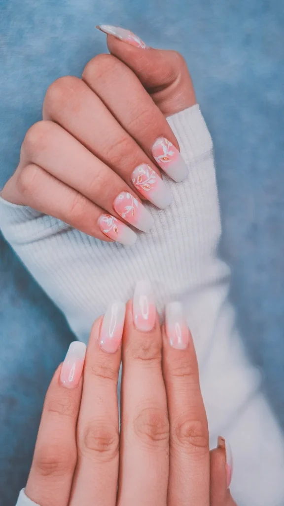 A close-up of long, square-shaped nails featuring a soft pink and milky white ombre gradient. The top hand showcases delicate white floral vine nail art with subtle gold leaf accents, while the bottom hand maintains a clean, ultra-glossy finish. The hands are posed against a white ribbed sweater and a textured blue background, embodying a sophisticated and minimalist "quiet luxury" aesthetic.