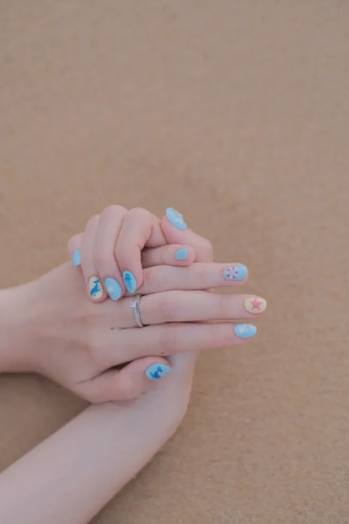 A close-up of a person's hands clasped together against a sandy beach background, showcasing vibrant Cancún nails. The manicure features light blue and sandy yellow nails adorned with ocean-themed nail art, including a pink starfish, a lifebuoy, a whale tail, and subtle sea waves.