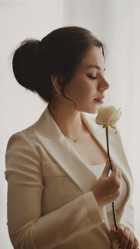 A profile view of a woman with dark hair styled in a polished, voluminous bun at the back of her head, with a soft, curled tendril framing her face. She wears a structured cream-colored blazer and a delicate necklace, holding a single white rose close to her face with her eyes closed in a serene expression. The lighting is soft and bright, emphasizing a clean and sophisticated aesthetic.