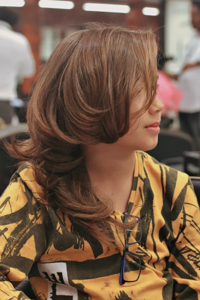 A side profile of a person with medium-to-long chestnut brown hair styled in a voluminous, layered blowout. The hair features significant lift at the roots and large, bouncy curls that flick inward and outward at the ends. One section of hair softly drapes over the side of the face. The person is wearing a yellow and black patterned shirt with a pair of glasses tucked into the neckline, seated in a brightly lit indoor setting with a blurred background.