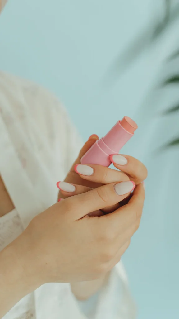 A close-up of hands with oval-shaped nails featuring a modern French manicure. The design uses a milky, sheer white base paired with vibrant, hot pink tips instead of traditional white. The hands are holding a small pink makeup stick against a soft blue background, emphasizing the flirty and coordinated color palette.