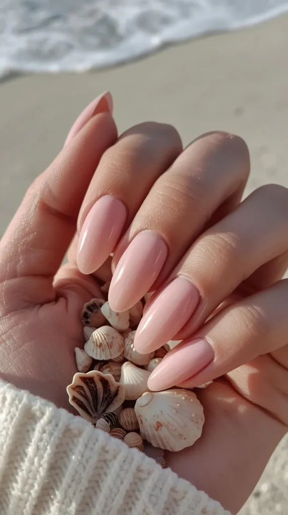 A close-up shot of a hand cupping a small handful of tiny, detailed seashells on a beach. The long, almond-shaped nails feature a glossy, soft milky-pink manicure. The sleeve of a cozy, cream-colored knit sweater is visible at the wrist. The background shows a soft-focus sandy shore with a hint of gentle white ocean foam in the distance.